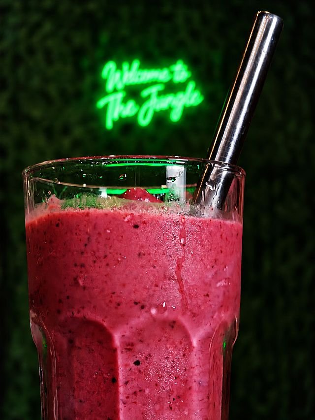 A close-up of a berry smoothie in a glass with a metal straw, with a neon "Welcome to The Jungle" sign in the background