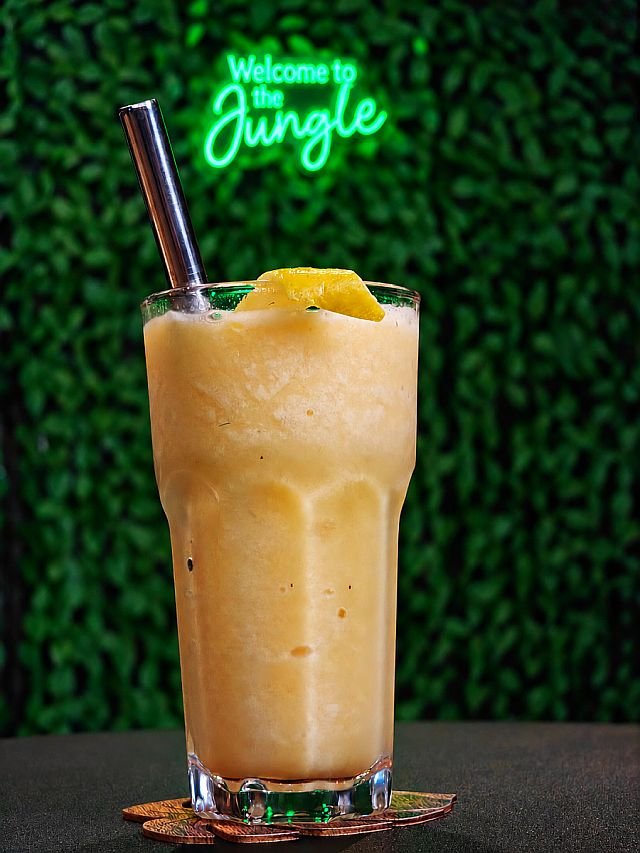 A frozen yellow smoothie with a metal straw and lemon garnish sits on a coaster in front of a neon "Welcome to the Jungle" sign