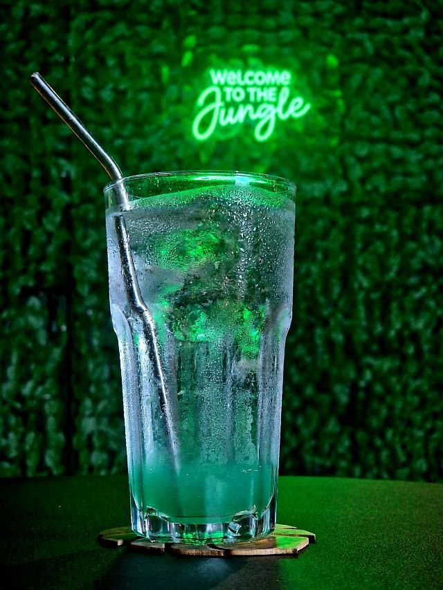 A tall glass of green cocktail with ice and a metal straw sits on a coaster in front of a neon "Welcome to the Jungle" sign