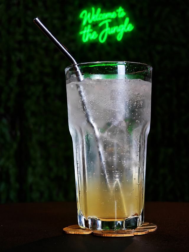 A tall glass of iced beverage with a metal straw sits on a wooden coaster in front of a green neon sign
