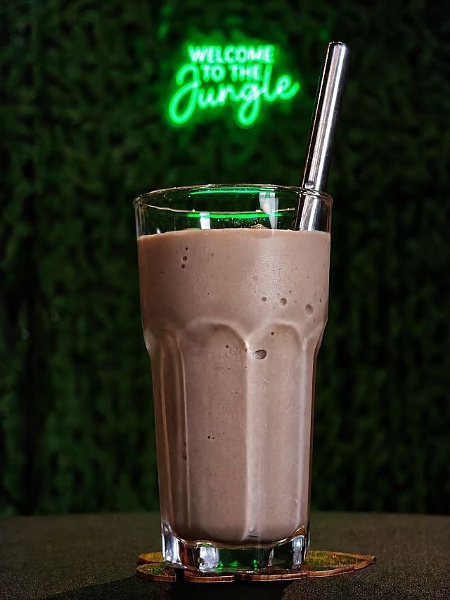 A chocolate milkshake with a metal straw sits on a leaf-shaped coaster in front of a neon "Welcome to the Jungle" sign
