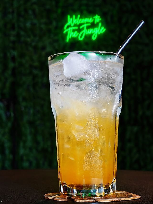 A tall glass of iced amber-colored drink with a metal straw sits on a wooden coaster in front of a green wall with a neon sign