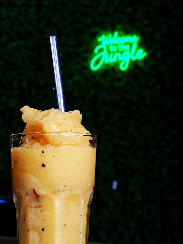 A tall glass of yellow passionfruit smoothie with a metal straw sits in front of a neon "Welcome to the Jungle" sign