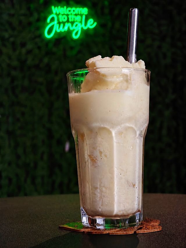 A tall glass of creamy, frozen pineapple drink with a metal straw sits on a coaster