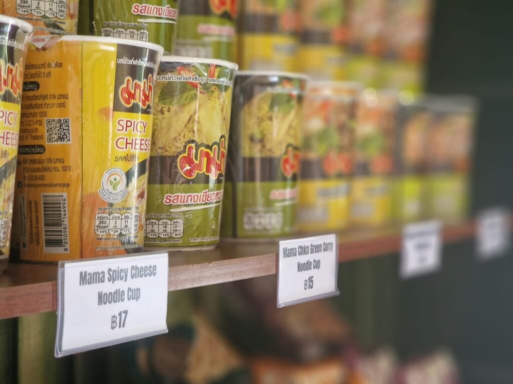 Mama Spicy Cheese and Chicken Green Curry noodle cups are displayed on a shelf with prices
