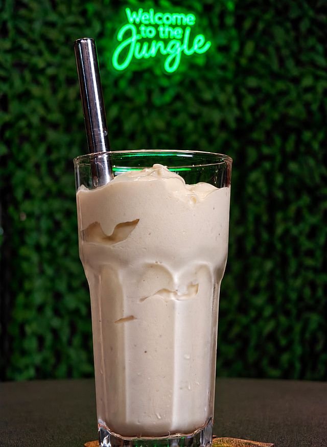 A creamy smoothie in a glass with a metal straw in front of a green leafy background and neon sign